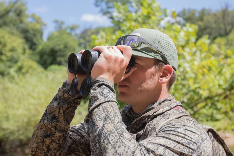 zeisssflinuse-810x540-1 A hunter looking through binoculars with a forest in the background.